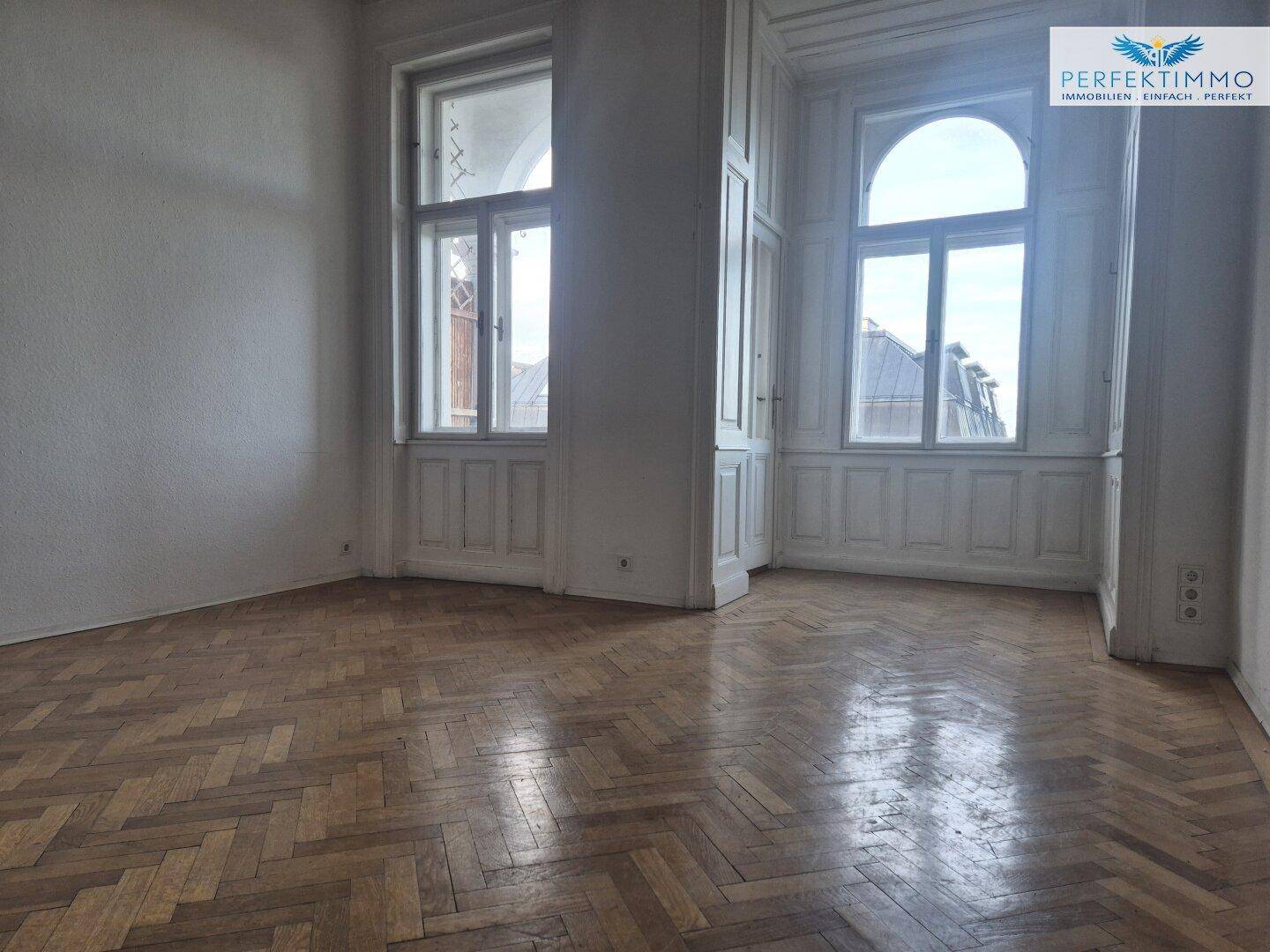 Heller Raum mit Fischgrätparkett und hohen Fenstern, die einen Blick auf die Umgebung ermöglichen.