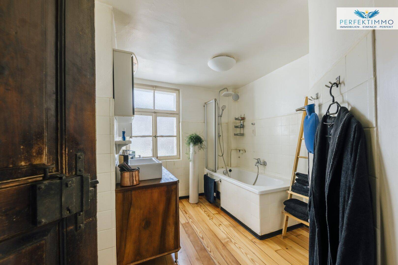 Helles Badezimmer mit Badewanne, Dusche und Holzfußboden, ausgestattet mit einem Fenster.