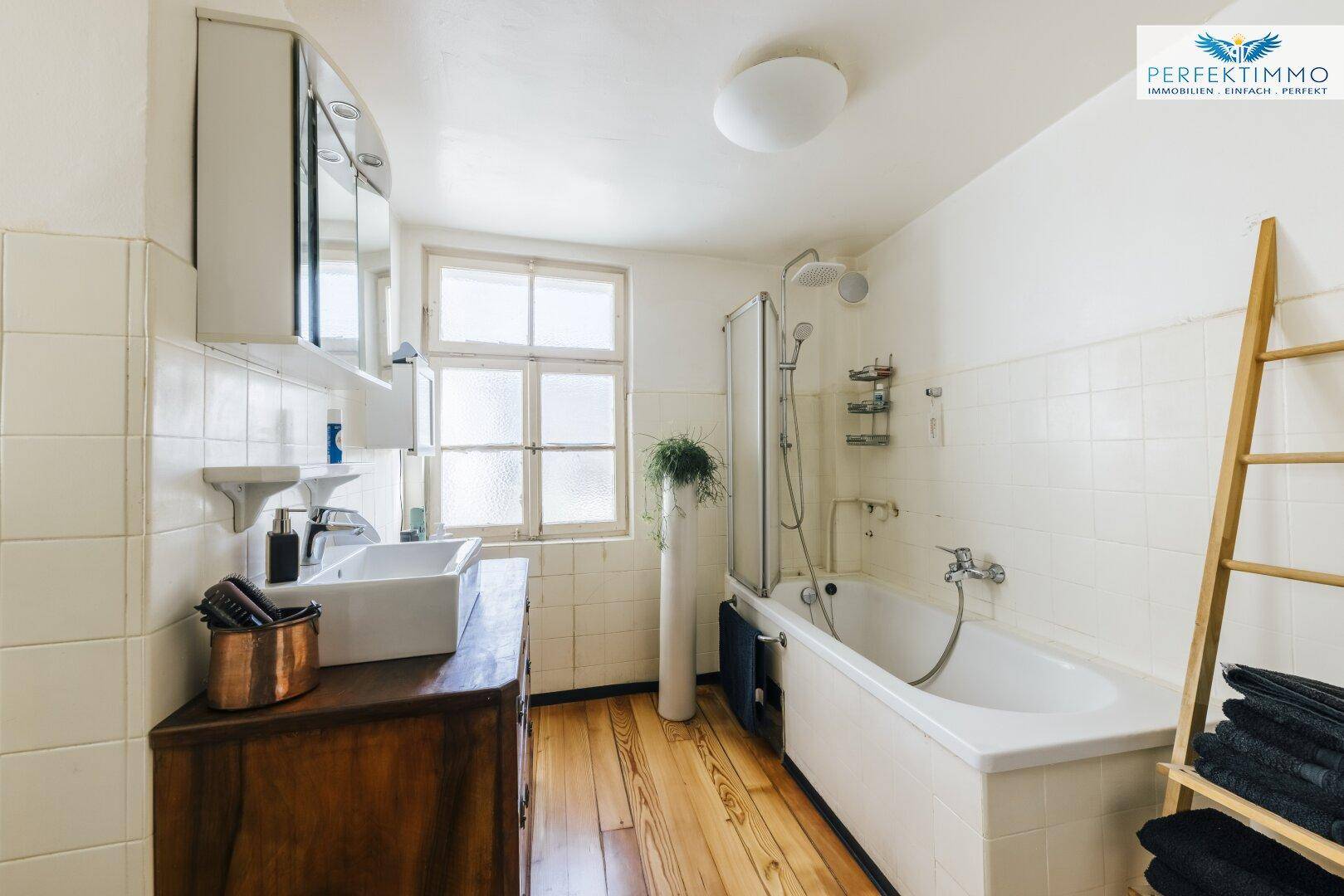 Funktionales Badezimmer mit Badewanne, Duschvorrichtung und Holzfußboden, hell gefliest.