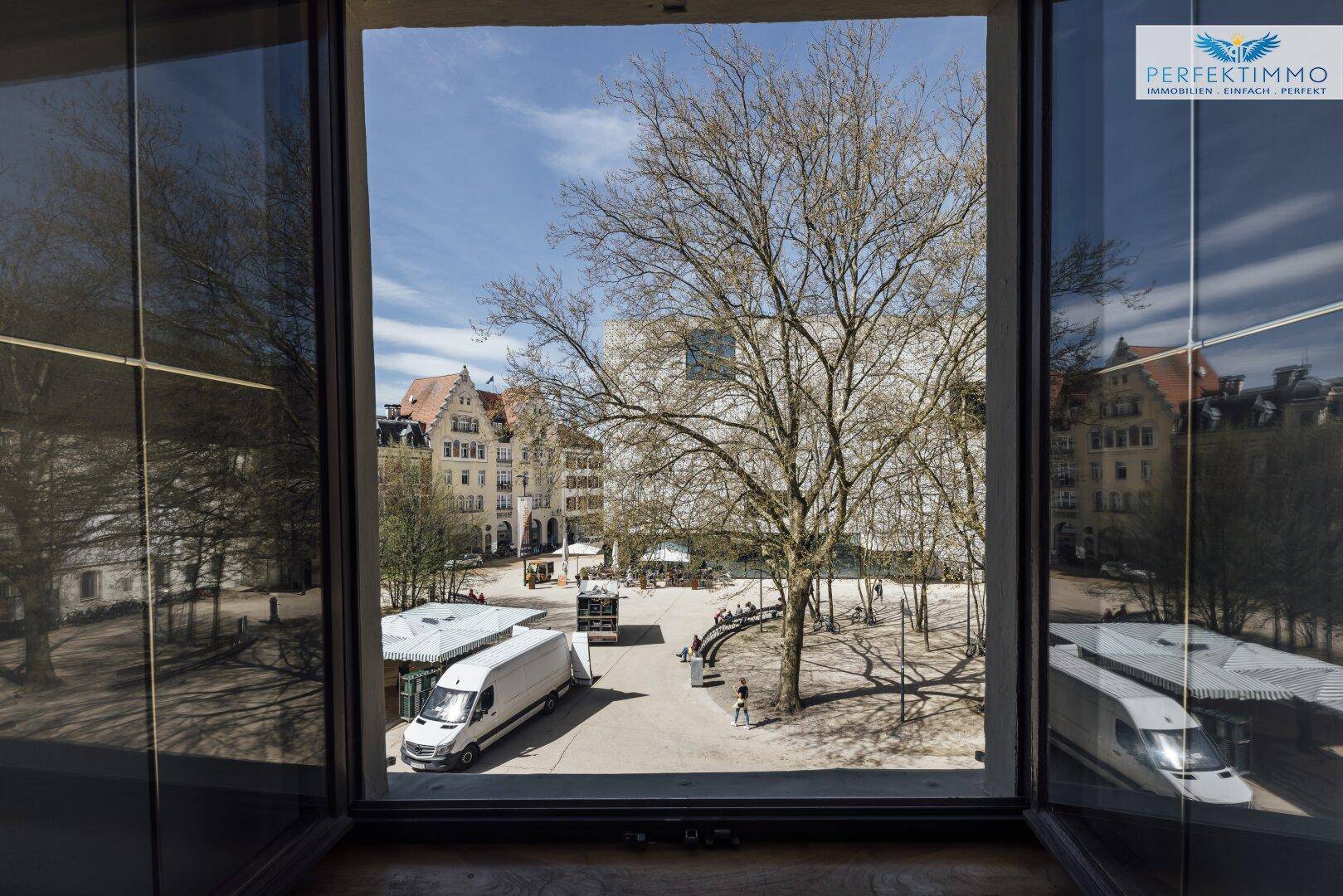 Blick aus dem Fenster auf einen belebten Stadtplatz mit Bäumen und Gebäuden.