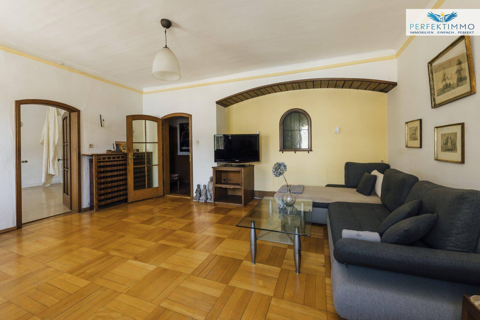 Helles Wohnzimmer mit Parkettboden, großem Sofa und Zugang zu weiteren Räumen.
