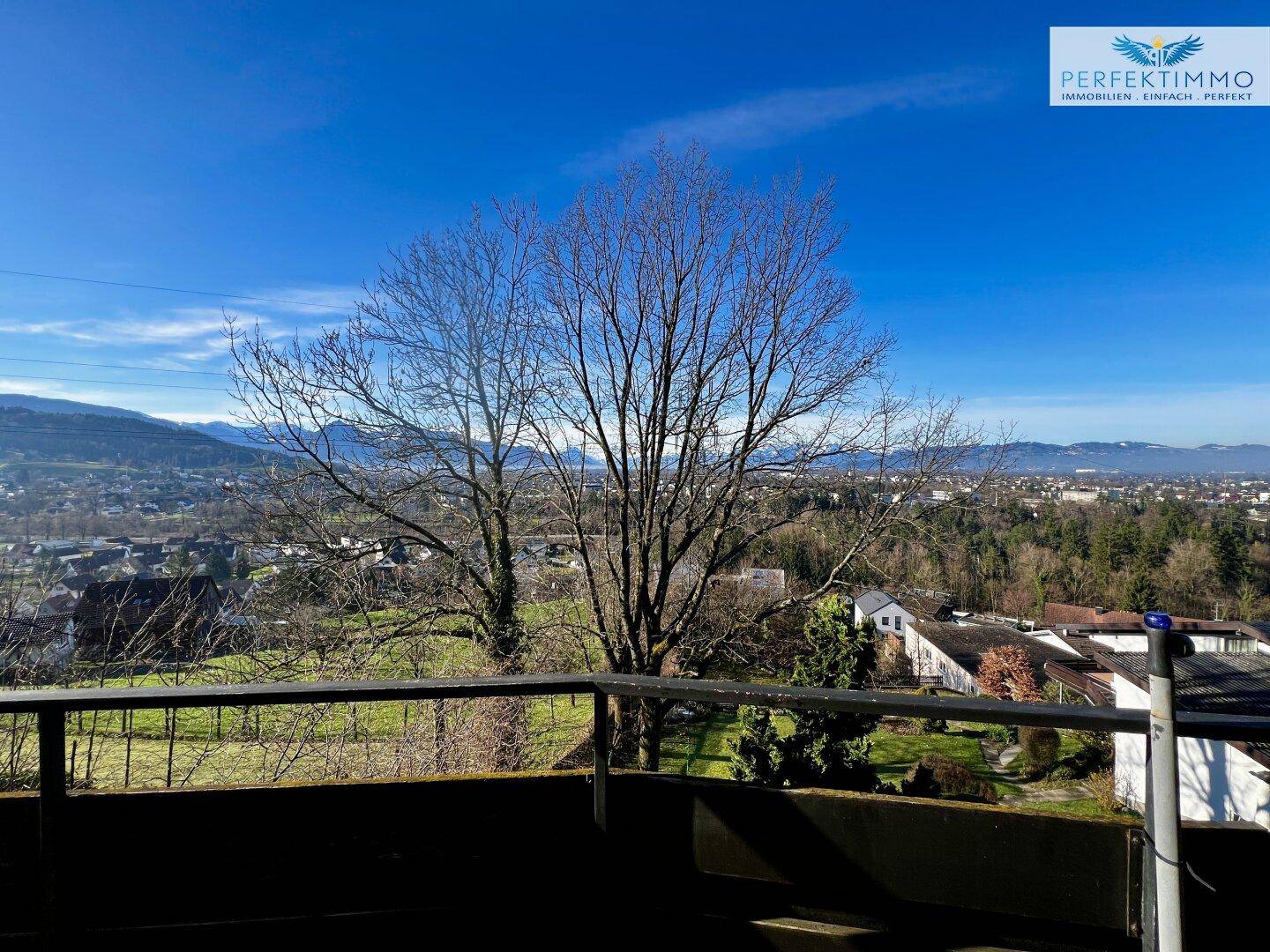 Weitblick von einem Balkon über die Stadt und die umliegende Berglandschaft an einem sonnigen Tag.