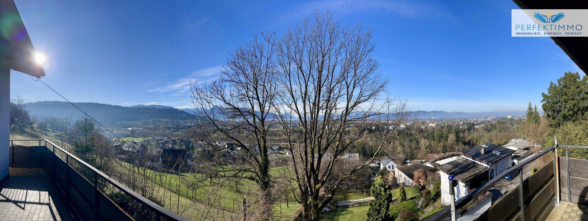 Breiter Panoramablick von einem Balkon über die hügelige Landschaft und die Stadt im Sonnenschein.