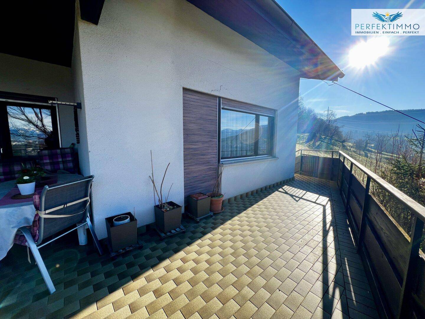 Sonniger Terrassenbereich mit Fliesenboden und Blick auf die grüne Umgebung und die Berge.