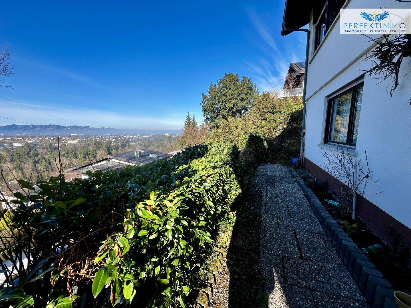 Schmaler Weg entlang der Hauswand, gesäumt von hohen, gepflegten Hecken.