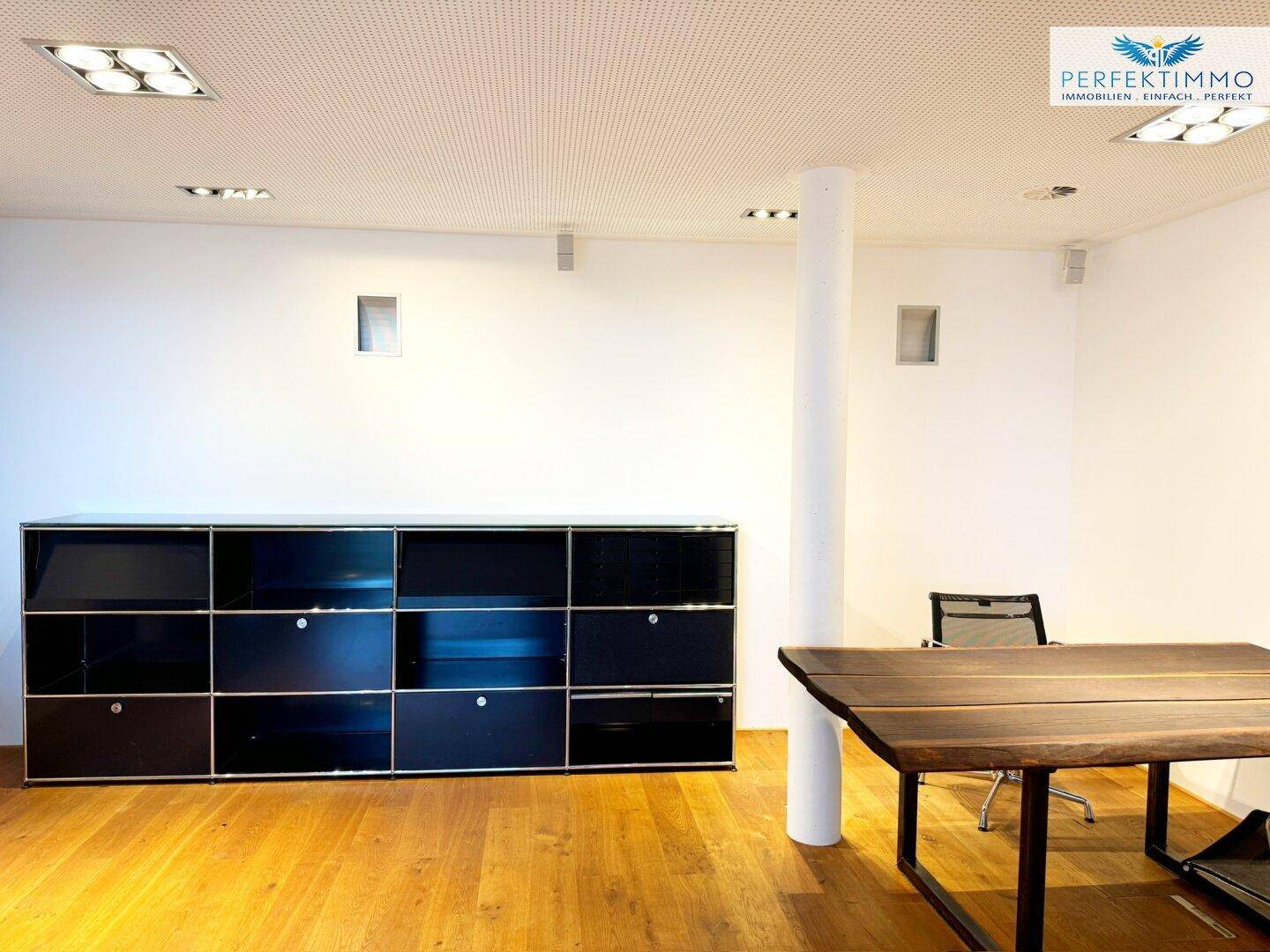 Gewerberaum mit schwarzem Regalsystem, Holztisch und weiß gestrichenen Wänden.