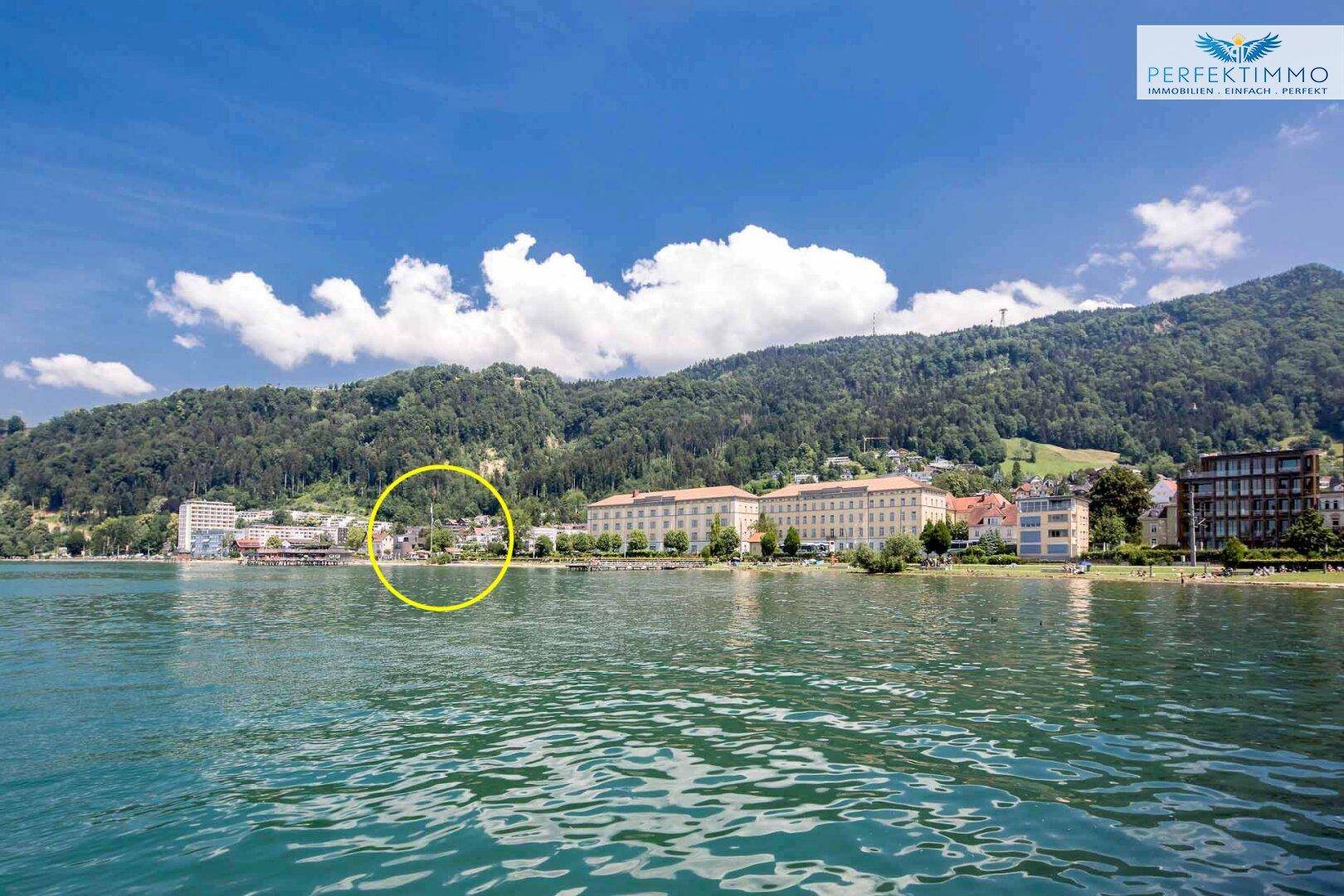 Panoramablick auf einen See mit bewaldeten Bergen und Gebäuden am Ufer.