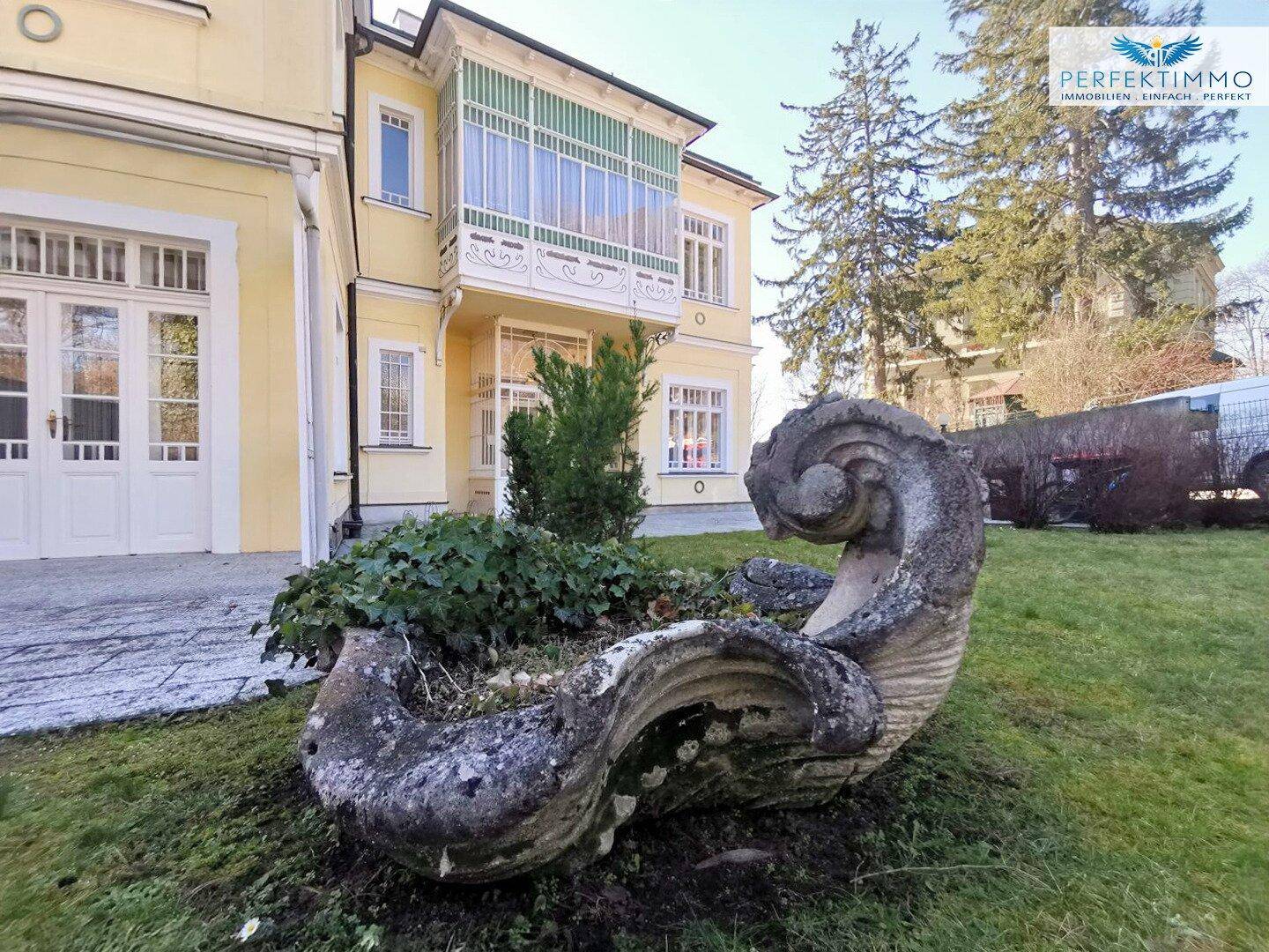 Gartenbereich mit Rasenfläche und einer dekorativen Steinskulptur vor der Villa.