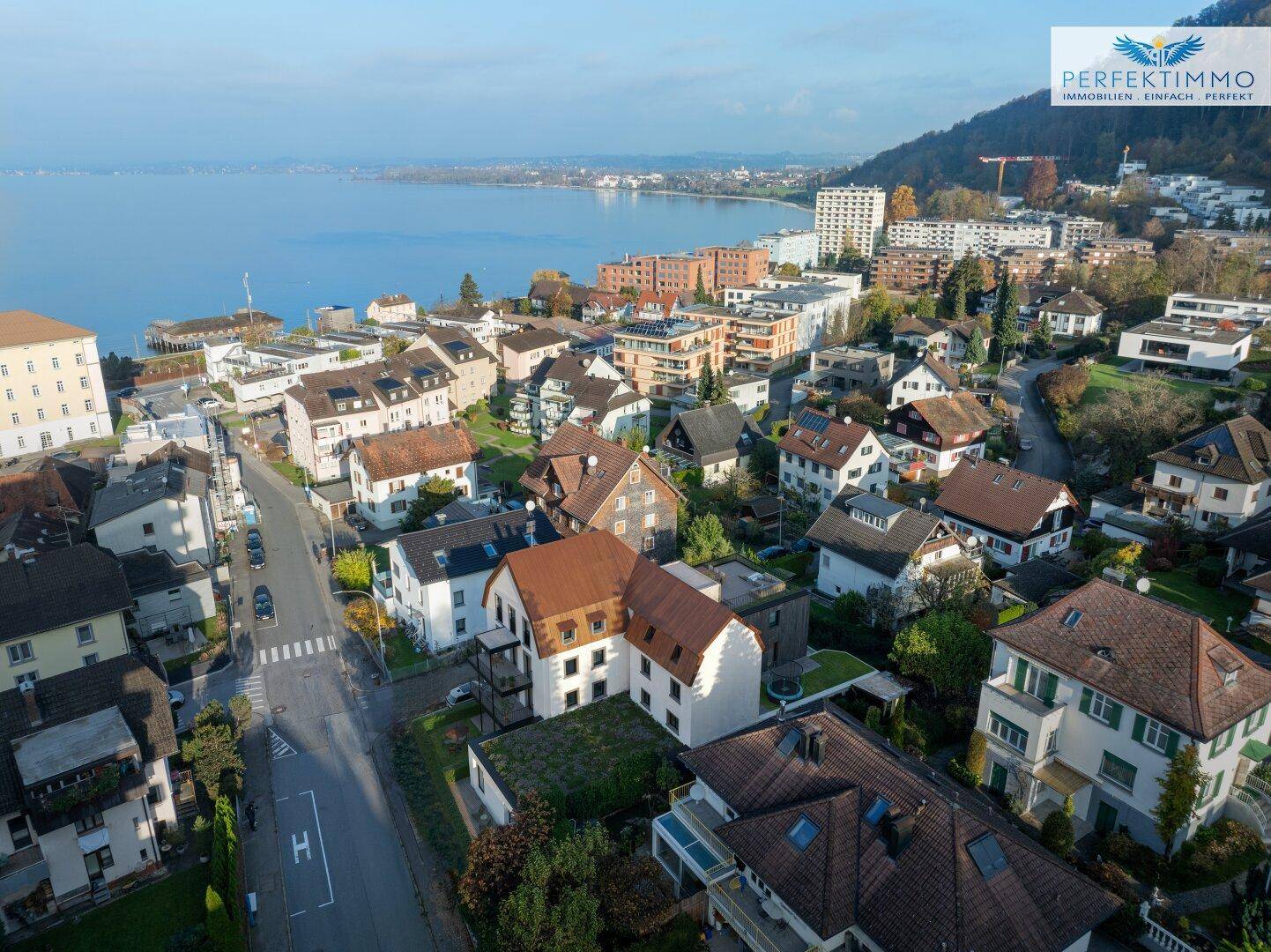 Weitläufige Luftaufnahme einer Stadt am Seeufer mit umliegenden Hügeln und Wohngebäuden.