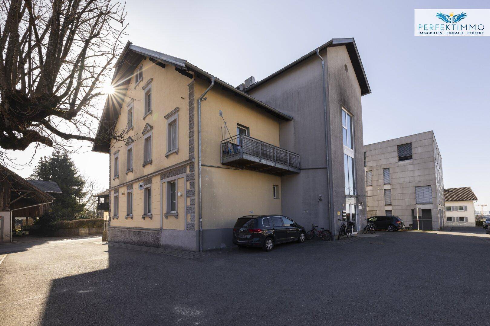 Seitenansicht des Gebäudes mit großem Fensterbereich, Balkon und angrenzendem Parkplatz.