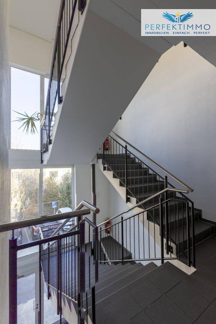 Helles Treppenhaus mit großen Fenstern, die viel Tageslicht hereinlassen, und robustem Geländer.