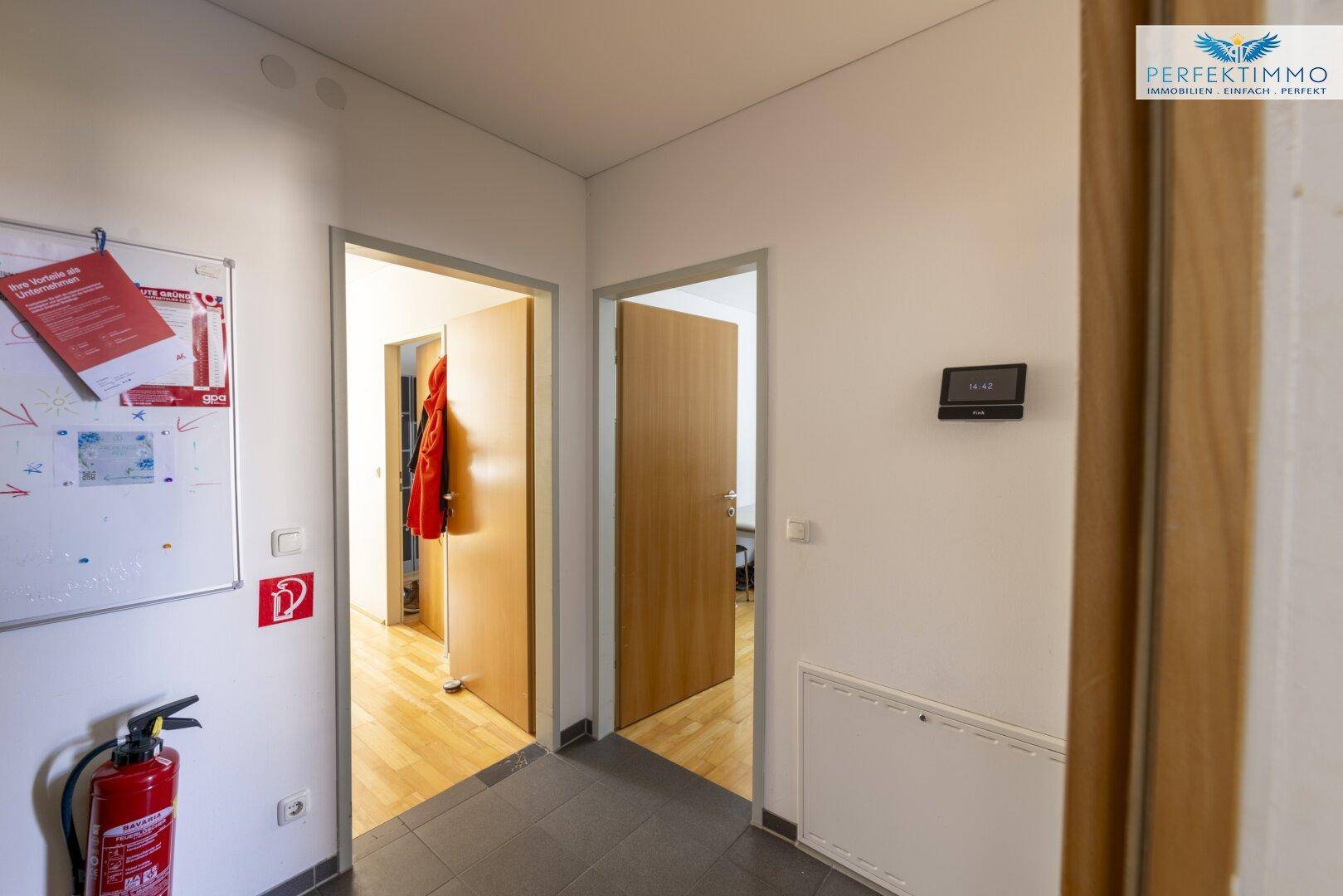 Heller Flur mit mehreren Türen, Fliesenboden und einem Feuerlöscher an der Wand.
