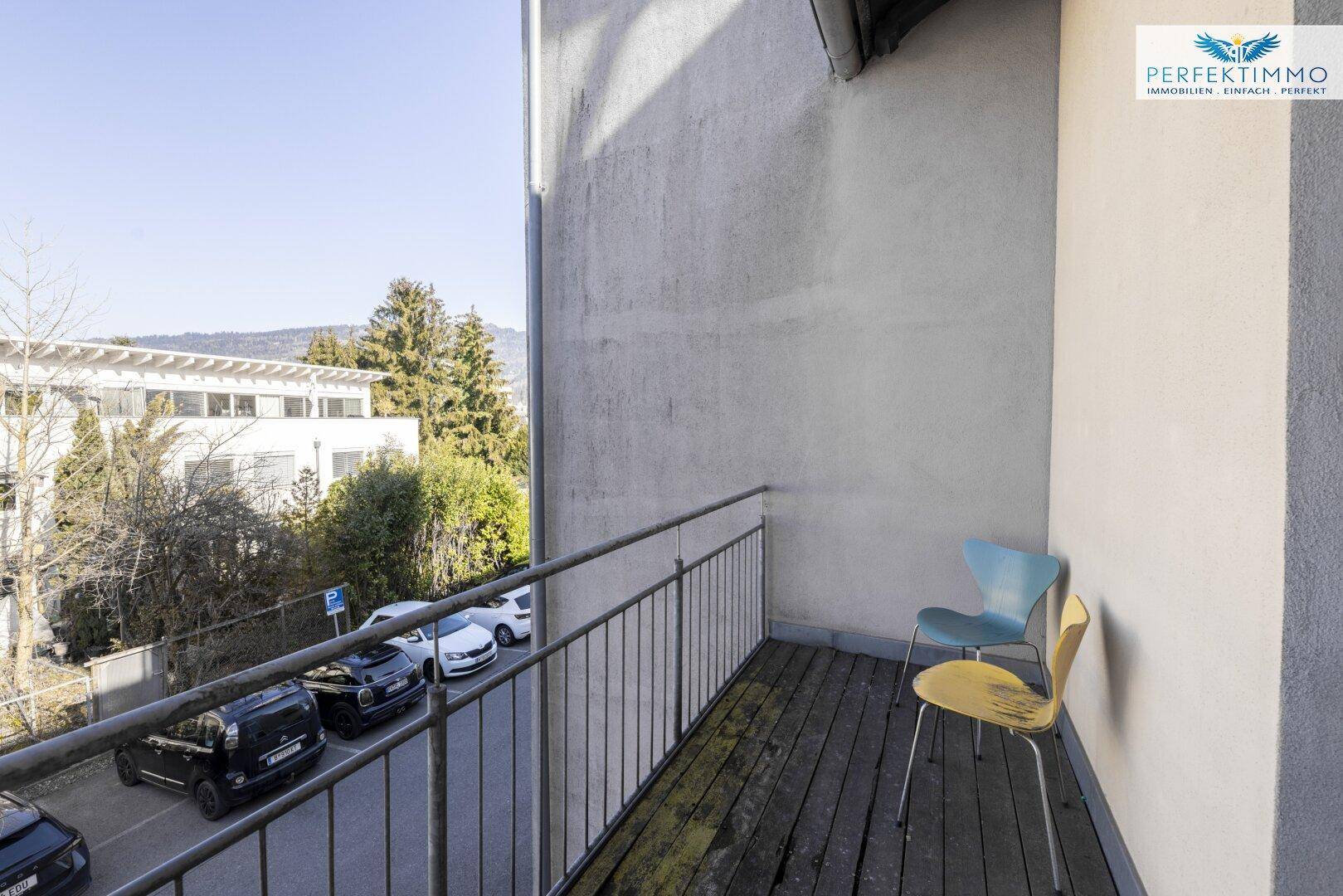 Kleiner Balkon mit Holzboden und Metallgeländer, möbliert mit zwei Stühlen und Blick auf die Umgebung.