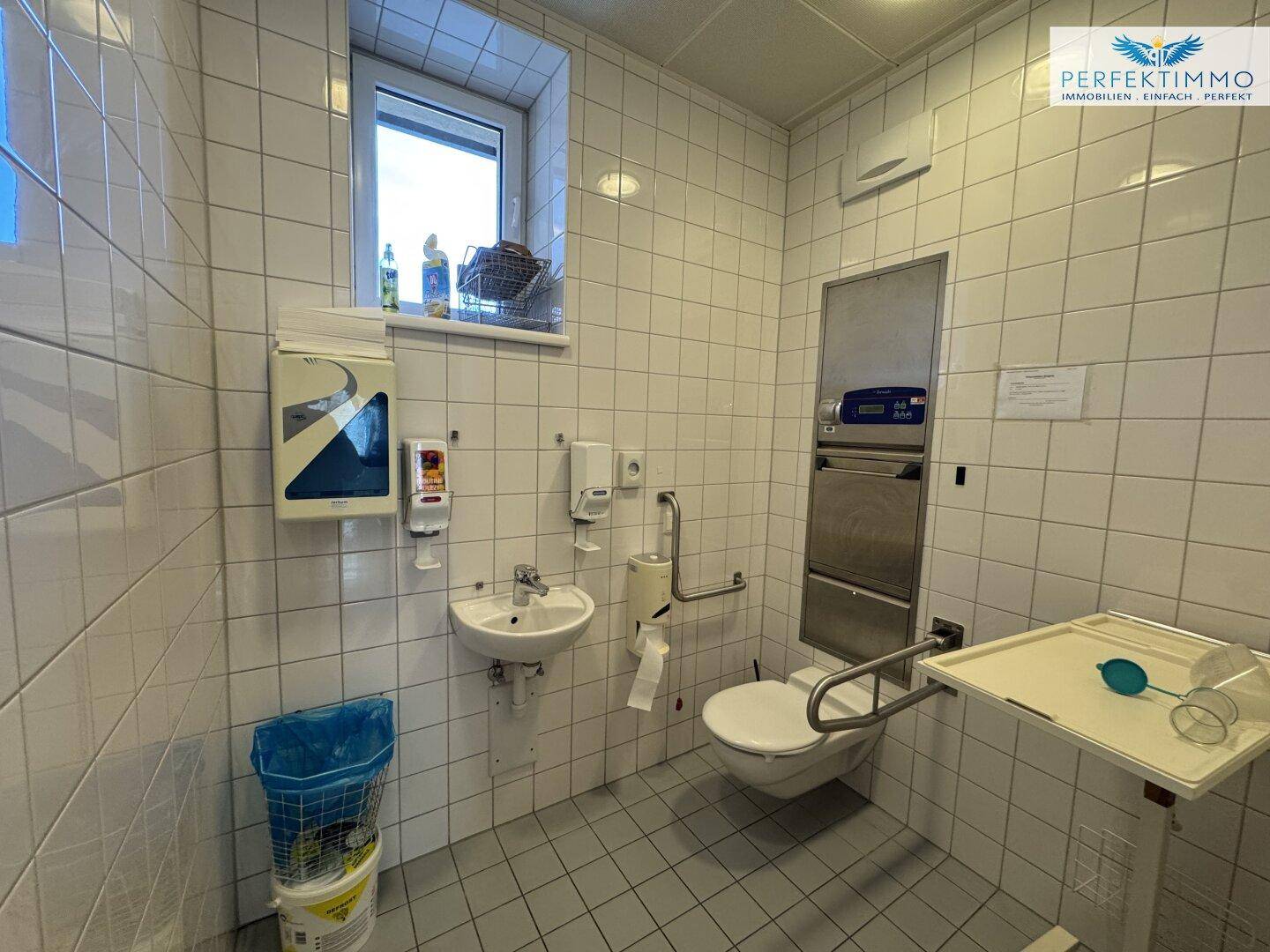 Barrierefreie Toilette mit Haltegriffen, Waschbecken, Wand-WC und Fenster für Tageslicht.