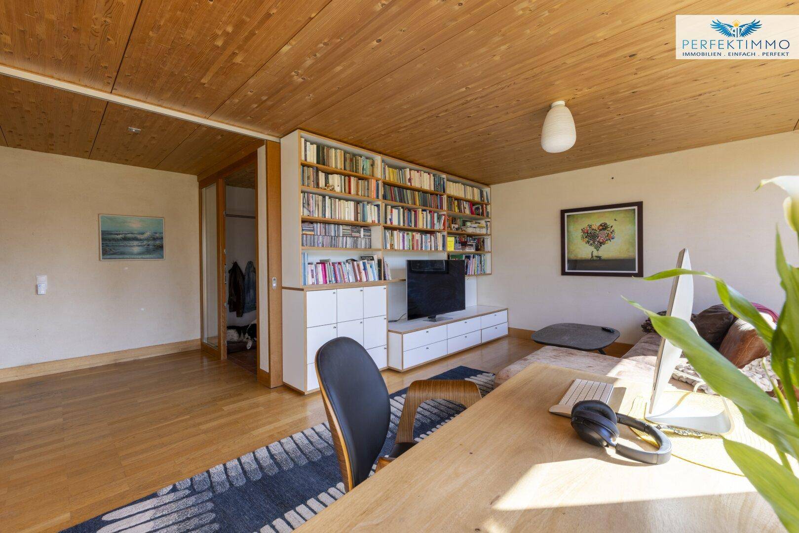 Geräumiges Wohnzimmer mit Bücherregal, Holzboden und großen Fenstern.