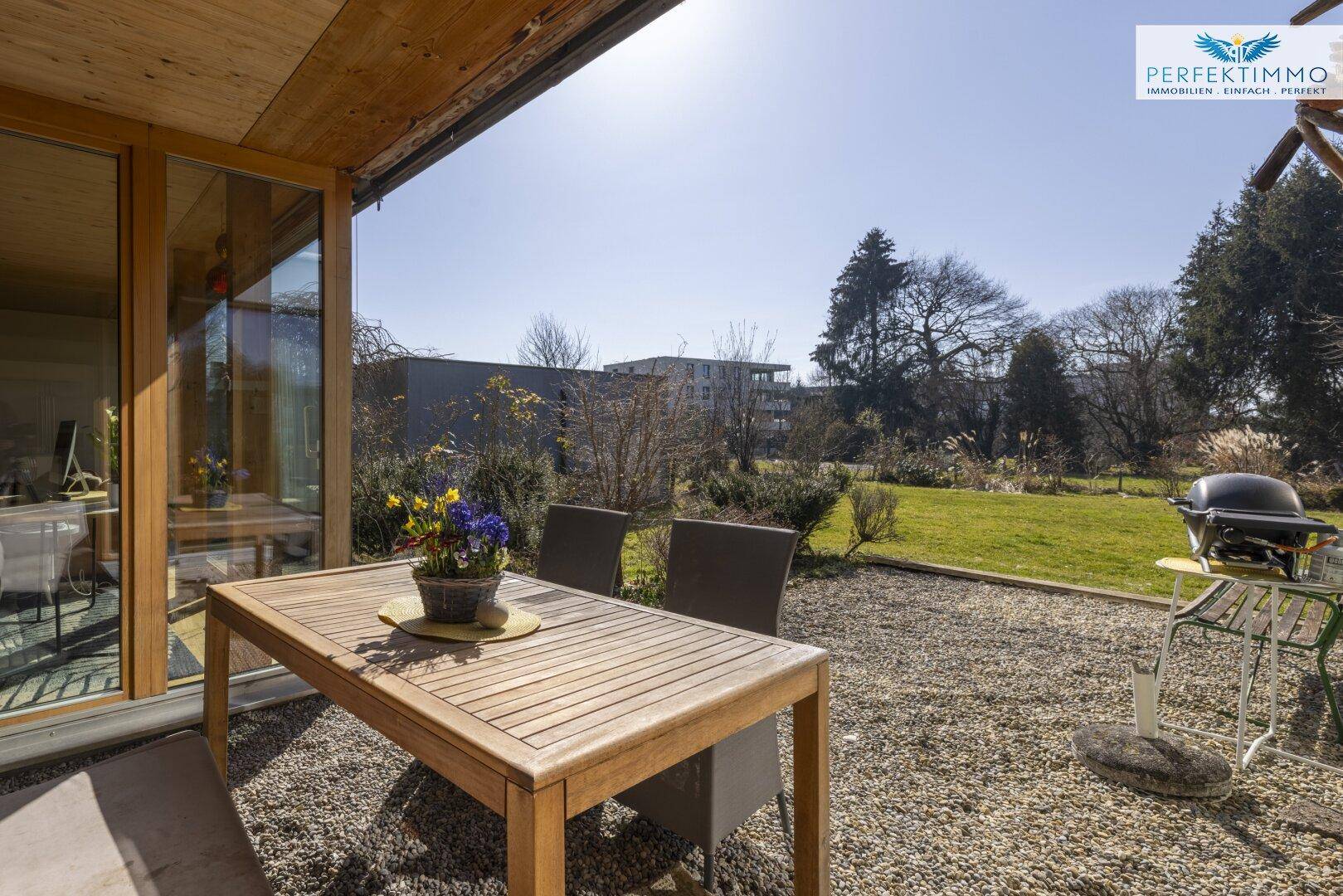 Sonniger Außenbereich mit Holztisch, Stühlen und Grillplatz im Garten.