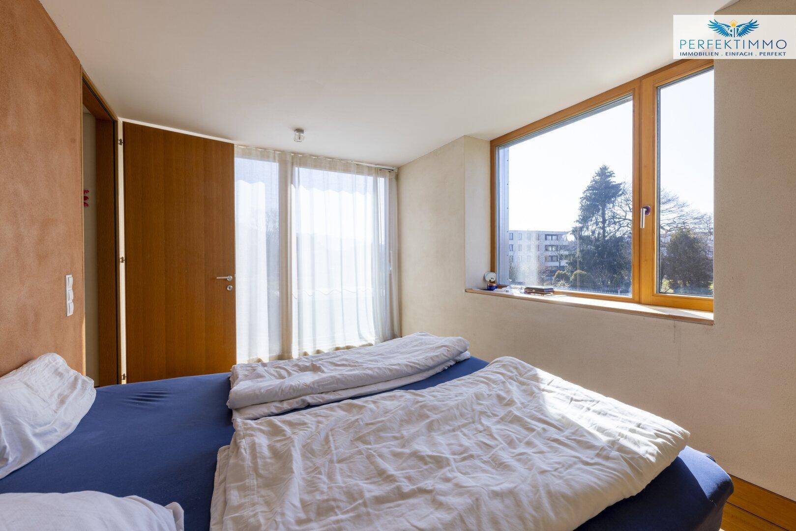 Schlafzimmer mit großem Fenster, Holzboden und viel natürlichem Licht.