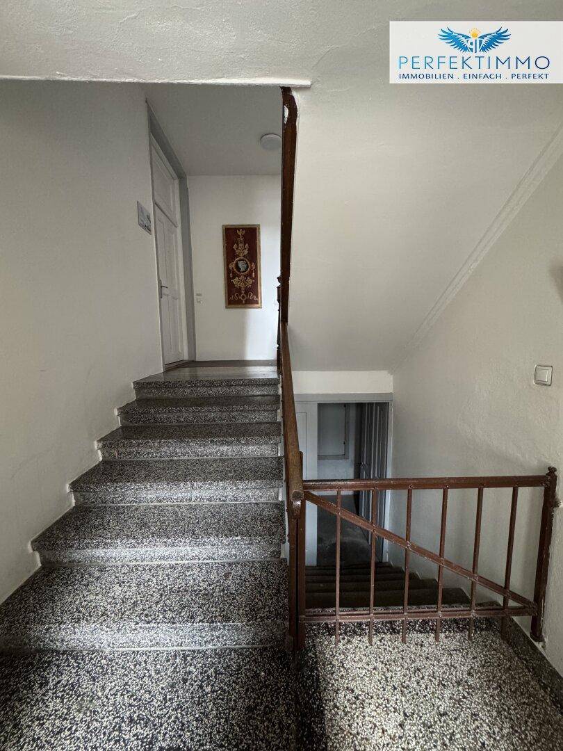 Treppenhaus mit dunklem Terrazzoboden und klassischem Holzgeländer in einem Mehrfamilienhaus.