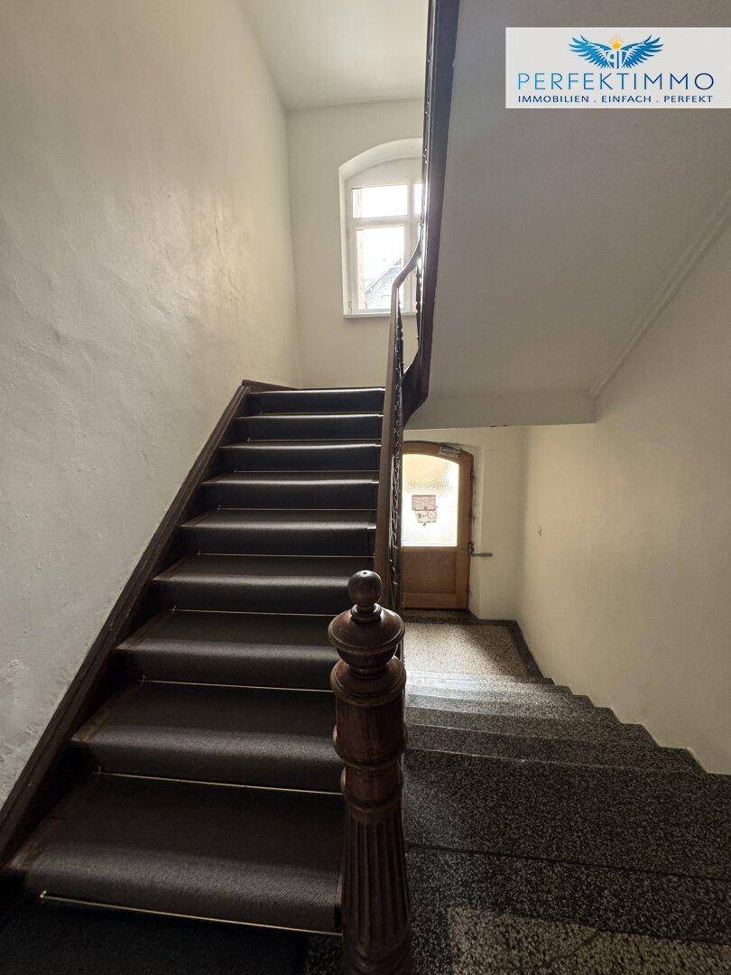 Helles Treppenhaus mit Fenster und dunklem Teppich auf den Stufen sowie einem Holzgeländer.