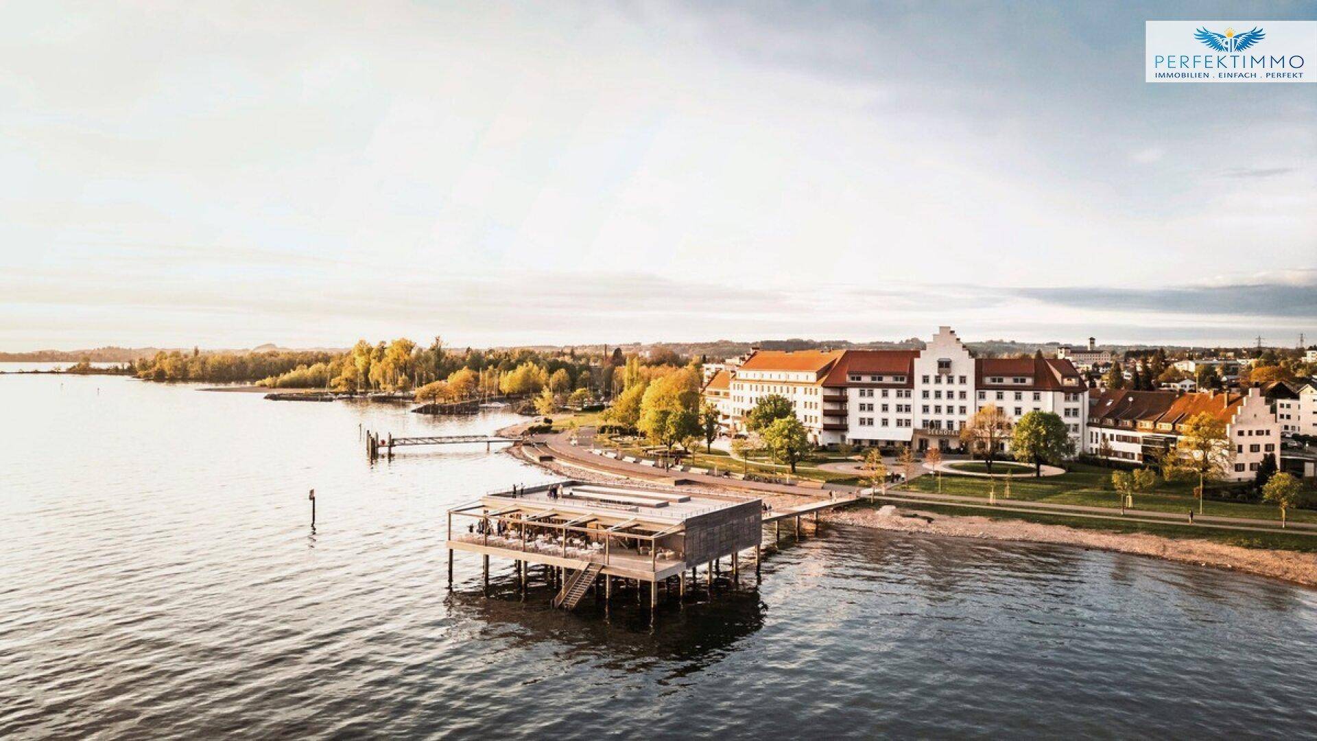 Panoramablick auf eine Seepromenade mit Gebäuden, einem Steg und einem Strandabschnitt bei Sonnenuntergang.