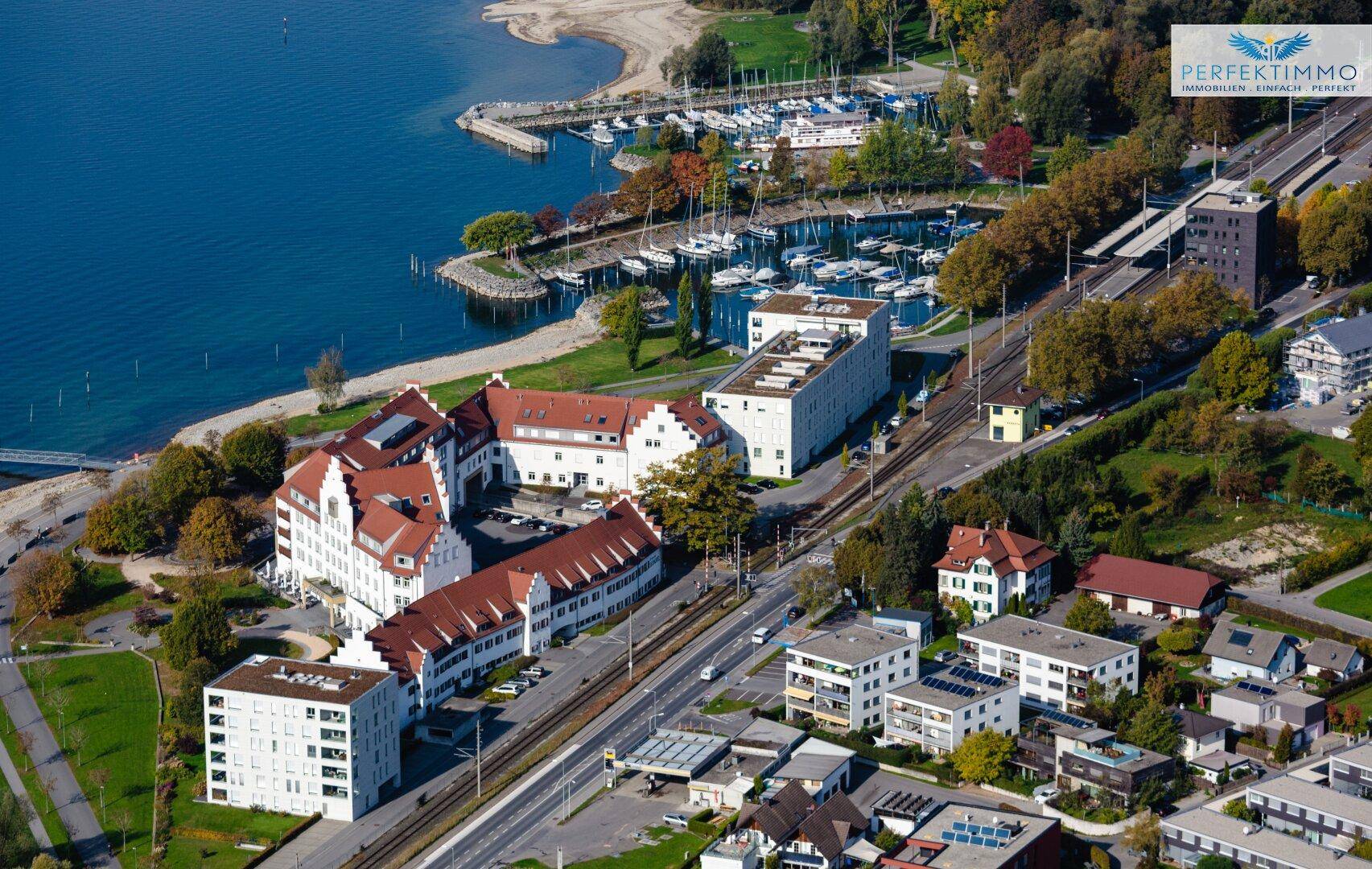 Vogelperspektive einer Küstenregion mit Gebäuden, einem belebten Yachthafen und angrenzenden Grünflächen.