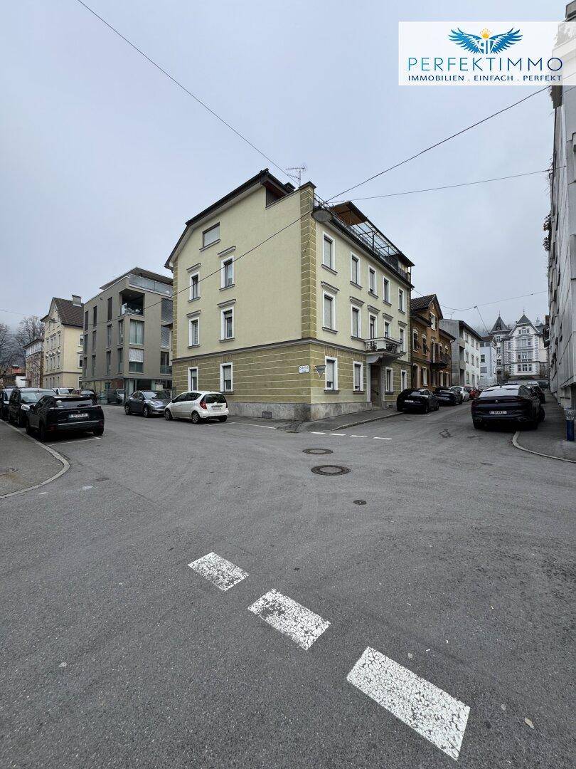 Ansicht des Mehrfamilienhauses von der Straßenecke mit parkenden Autos und umliegenden Gebäuden.