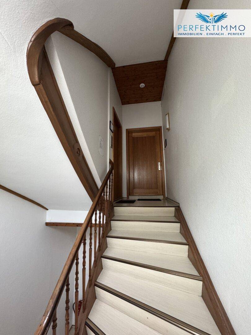 Treppenhaus mit Holzgeländer und einer Holztür am oberen Ende, die zu einer Wohnung führt.