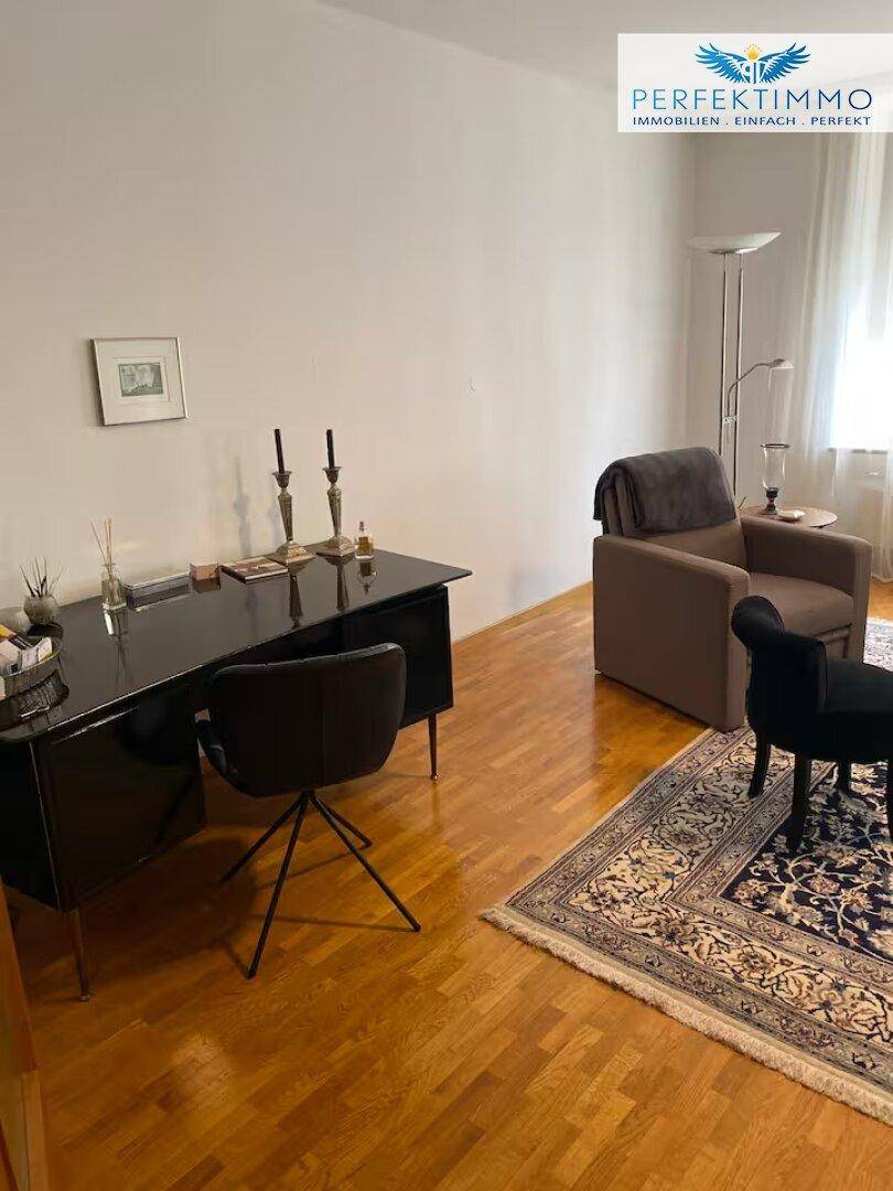 Arbeitsbereich im Wohnzimmer mit Schreibtisch, Sessel und Holzboden, beleuchtet durch Tageslicht.