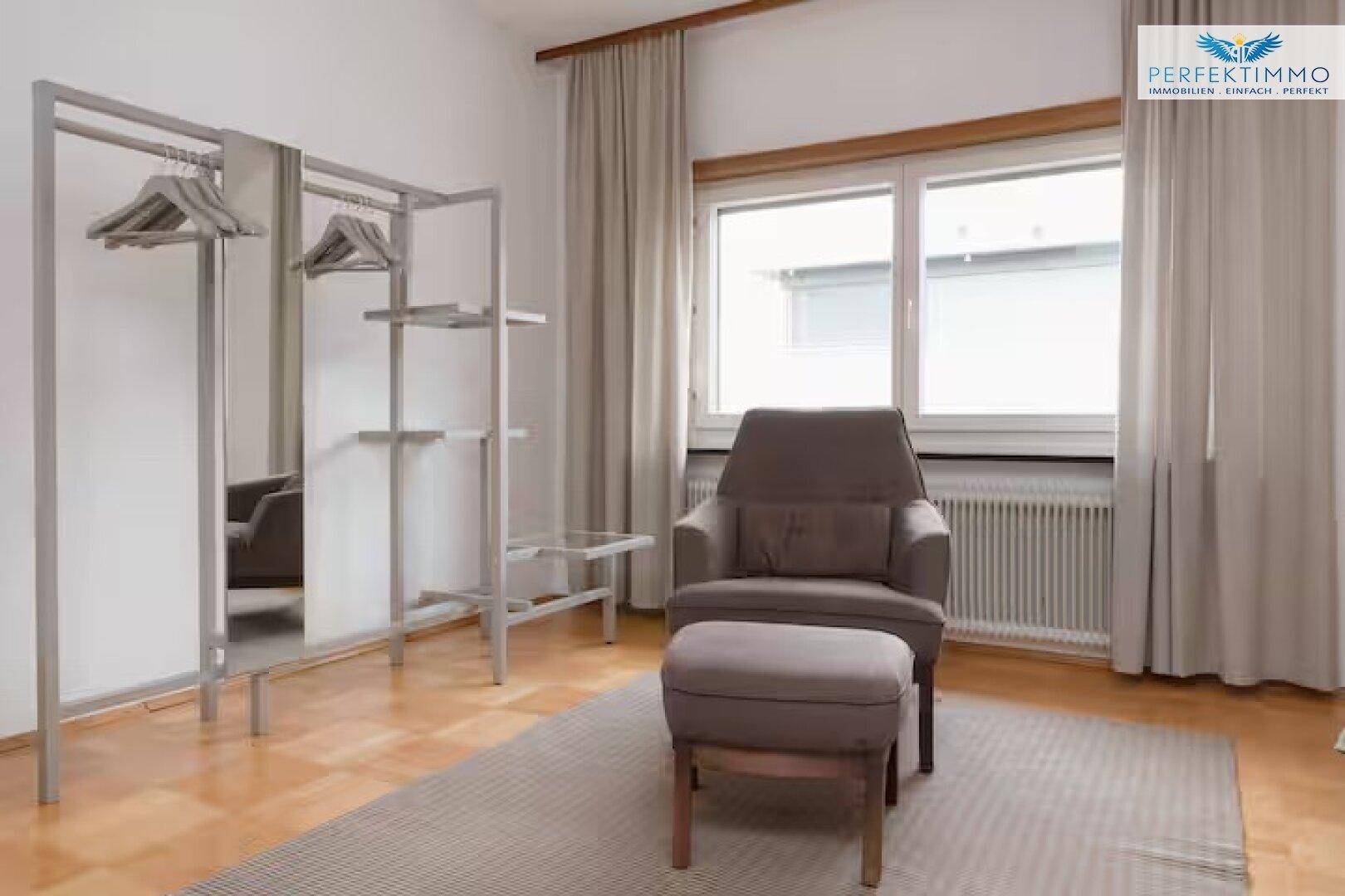 Schlafzimmer mit Fenster, Sessel, Fußhocker und offenem Kleiderständer.