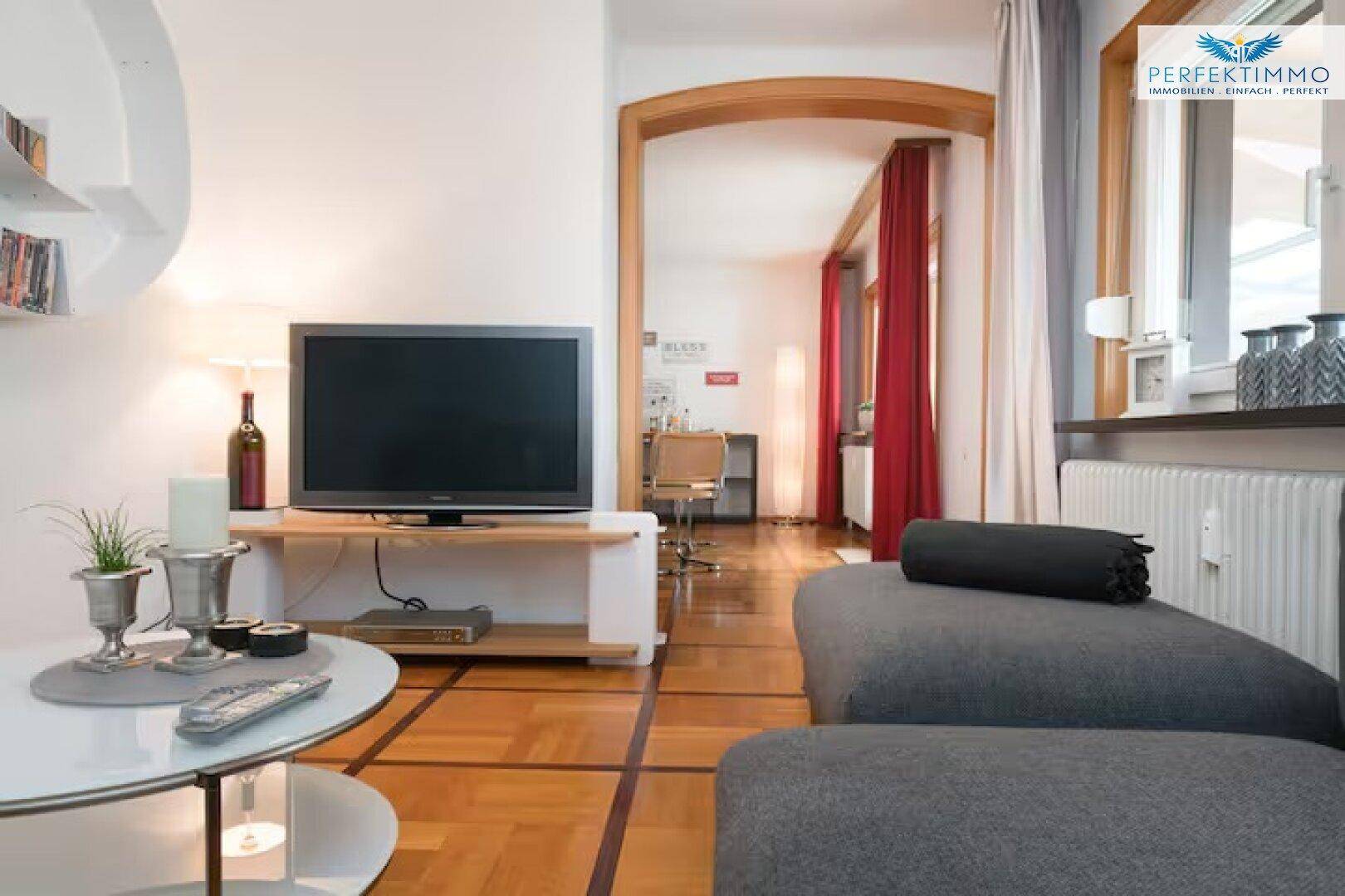 Wohnzimmer mit Sofa, Fernseher und Parkettboden, verbunden mit dem Essbereich.