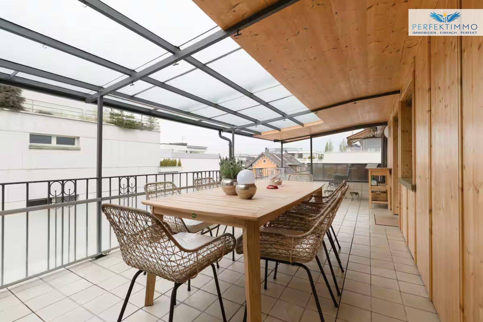 Große überdachte Terrasse mit Holzwänden, Esstisch und Blick auf die umliegenden Gebäude.