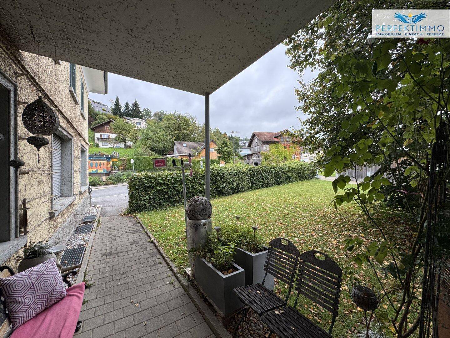 Außenbereich mit gepflasterter Terrasse, Rasenfläche und Häusern im Hintergrund.