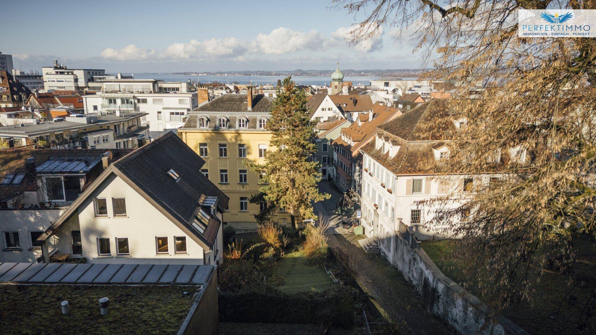 Weitläufiger Blick über die Stadt mit Wohngebäuden, einem See und bewaldeten Hügeln.