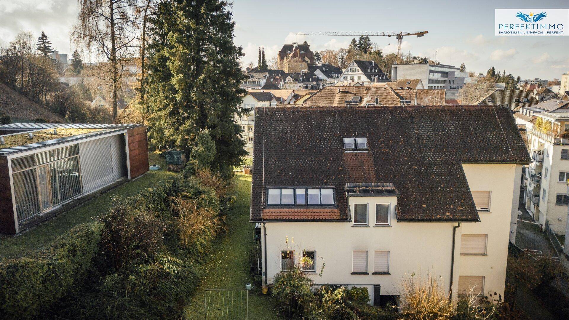 Blick über Wohngebäude und Gärten mit einem Baukran in der Ferne, umgeben von Bäumen.