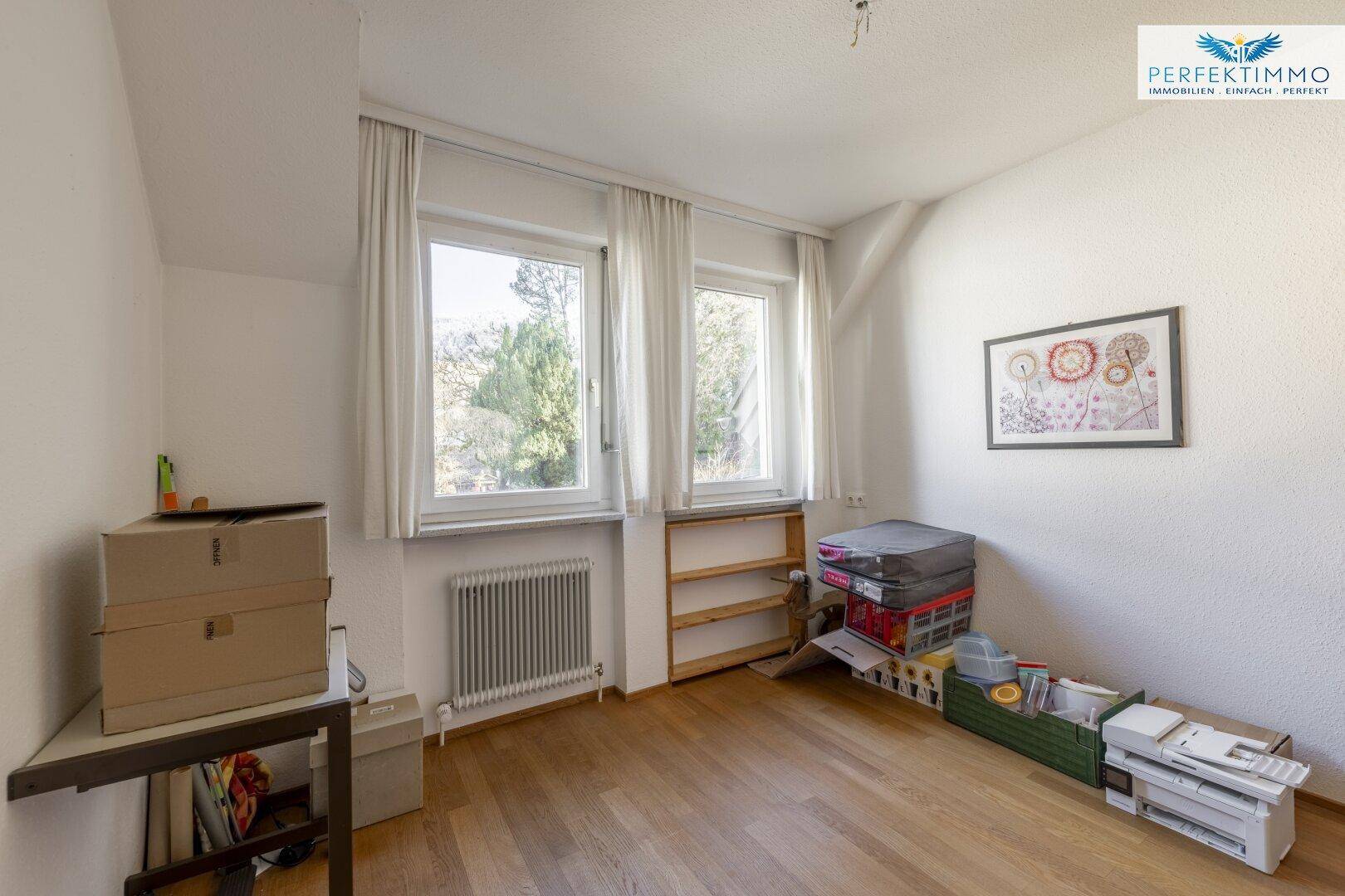 Helles Zimmer mit Holzboden, zwei Fenstern und einem Heizkörper, derzeit als Abstellraum genutzt.