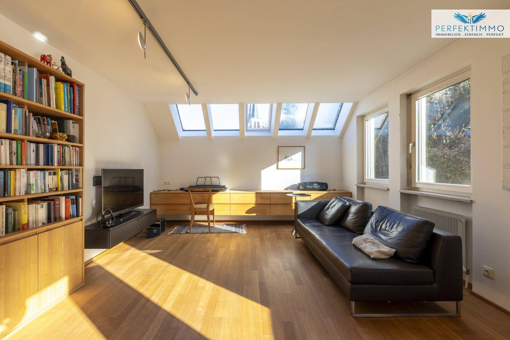 Heller Wohnbereich mit Sofa, niedrigem Schrank, Bücherregal und mehreren Dachfenstern.