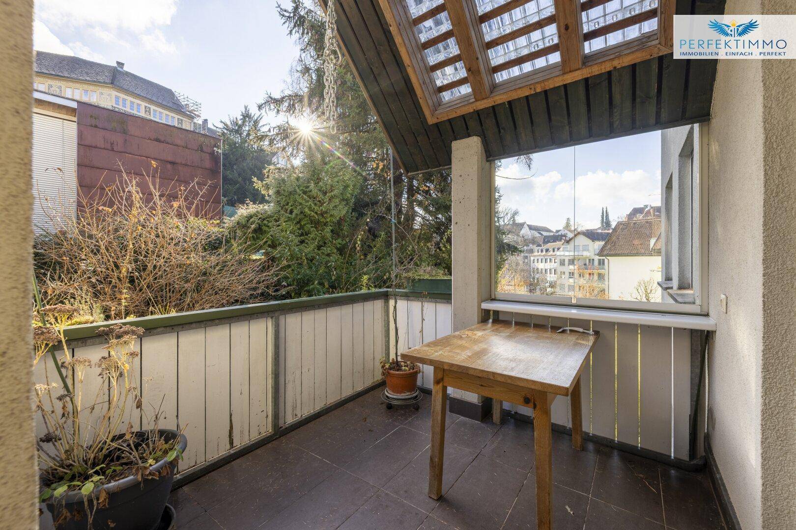Sonniger Balkon mit Holztisch, Fliesenboden und Blick auf die Stadt und umliegende Bäume.