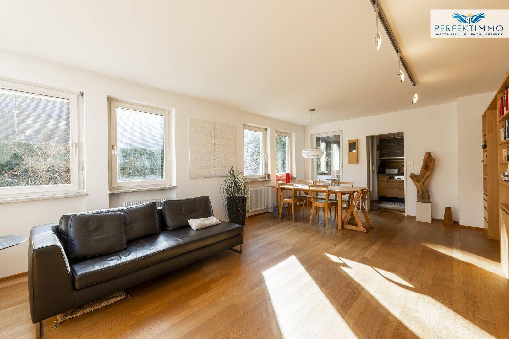 Lichtdurchfluteter Wohn-Essbereich mit bequemer Couch, Esstisch und mehreren Fenstern.