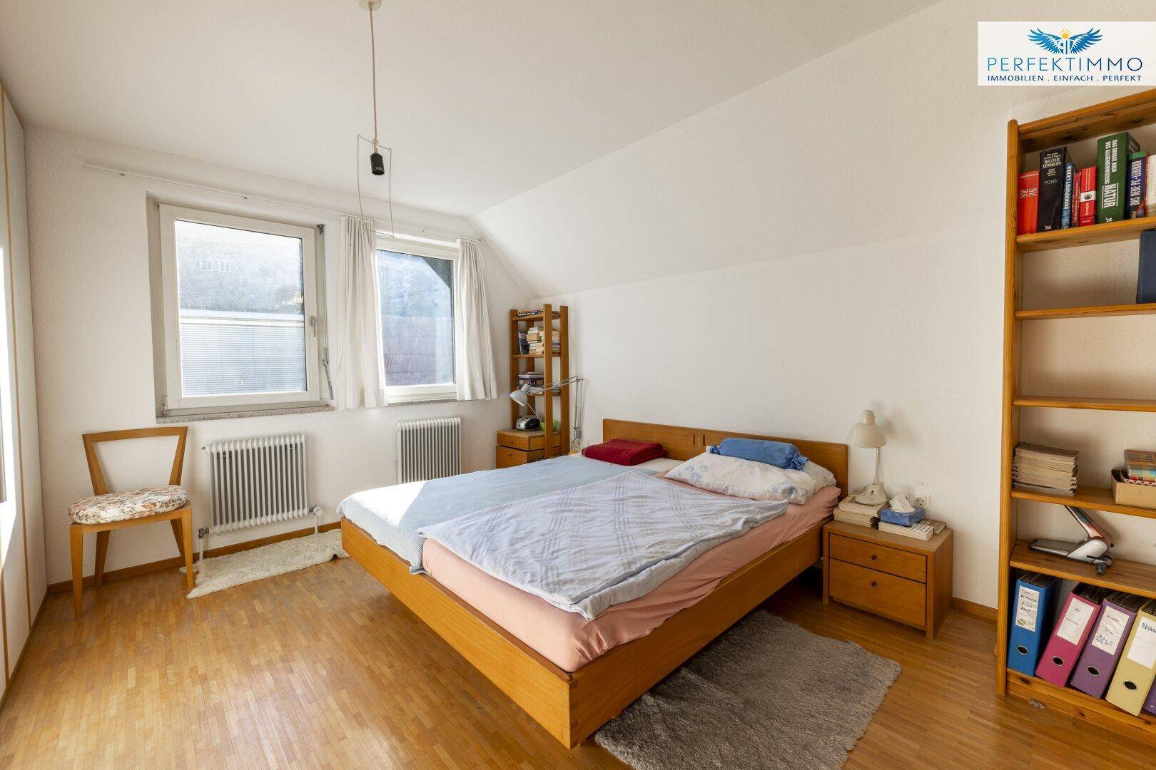 Gemütliches Schlafzimmer mit Holzbett, Bücherregal und zwei Fenstern, hell und einladend.