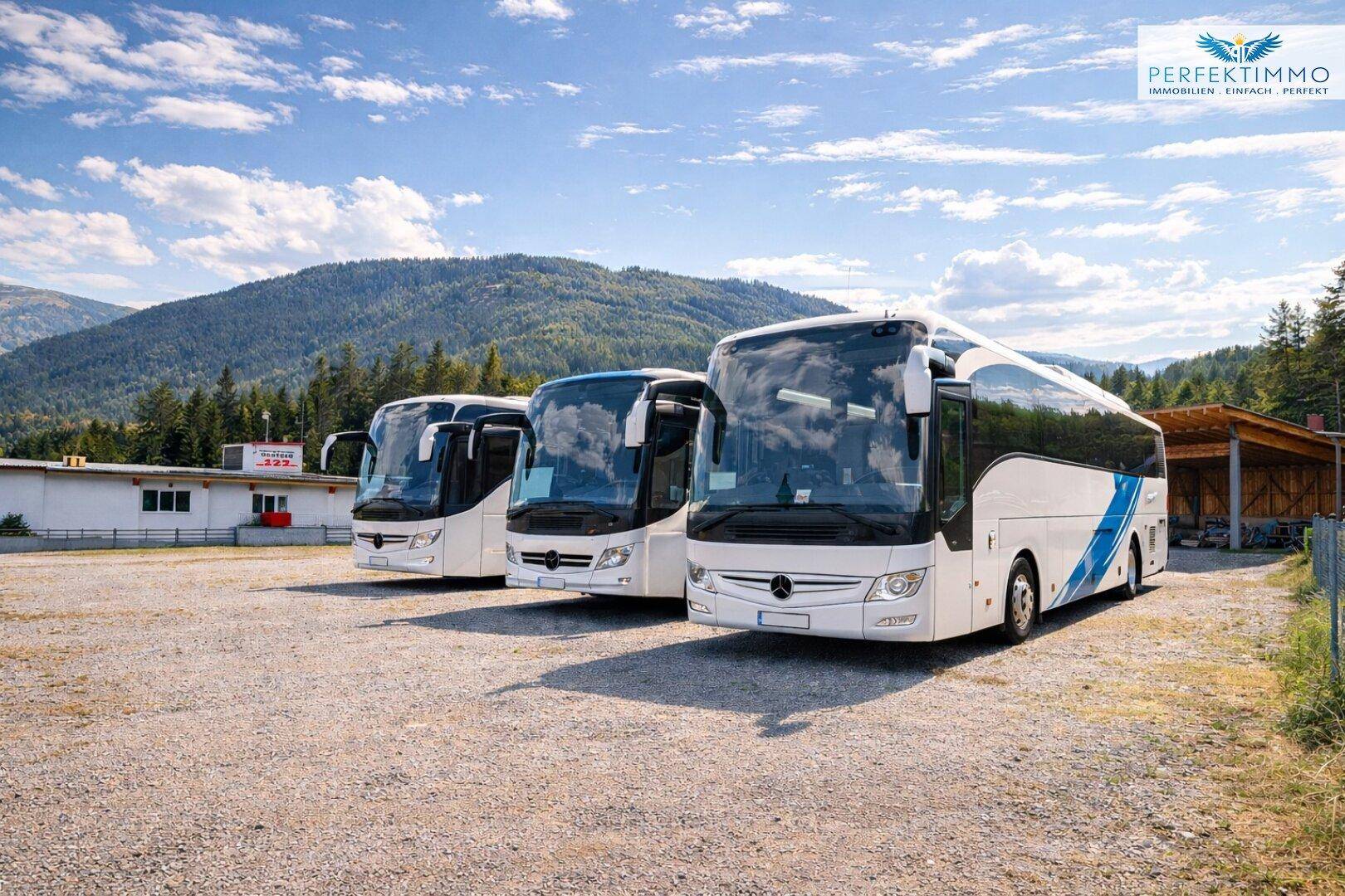 Drei Reisebusse parken auf einem Schotterplatz vor einem Gewerbegebäude mit Bergkulisse an einem sonnigen Tag.