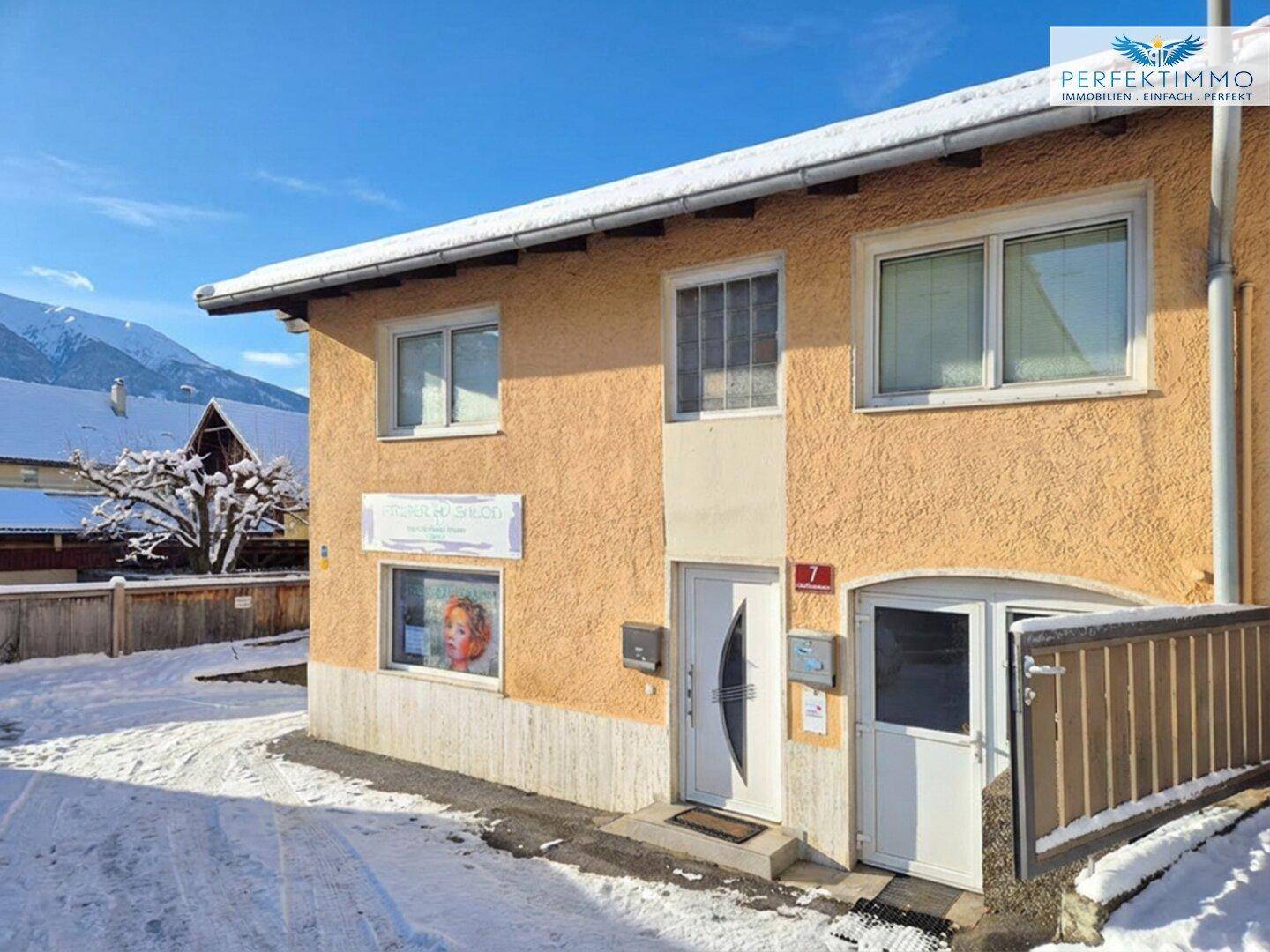 Gebäudeansicht im Winter mit Schnee auf dem Dach und den umliegenden Flächen, im Hintergrund Berge.