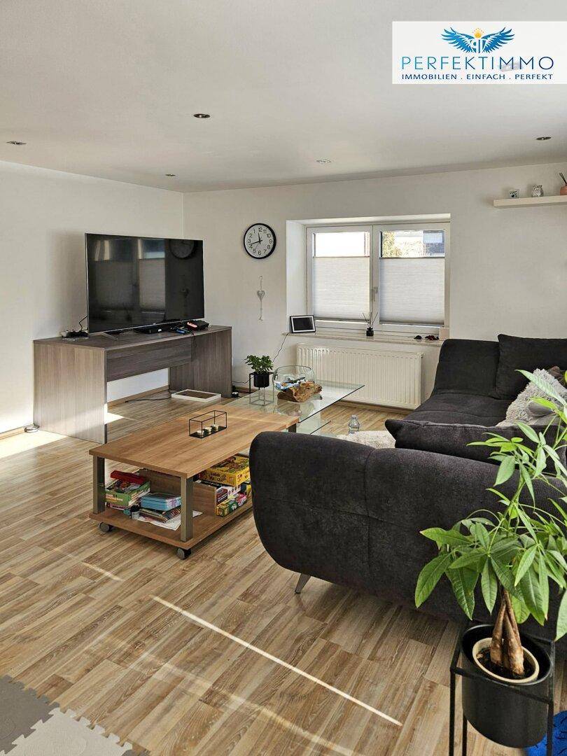 Gemütliches Wohnzimmer mit dunklem Ecksofa, Holztisch und Laminatboden.