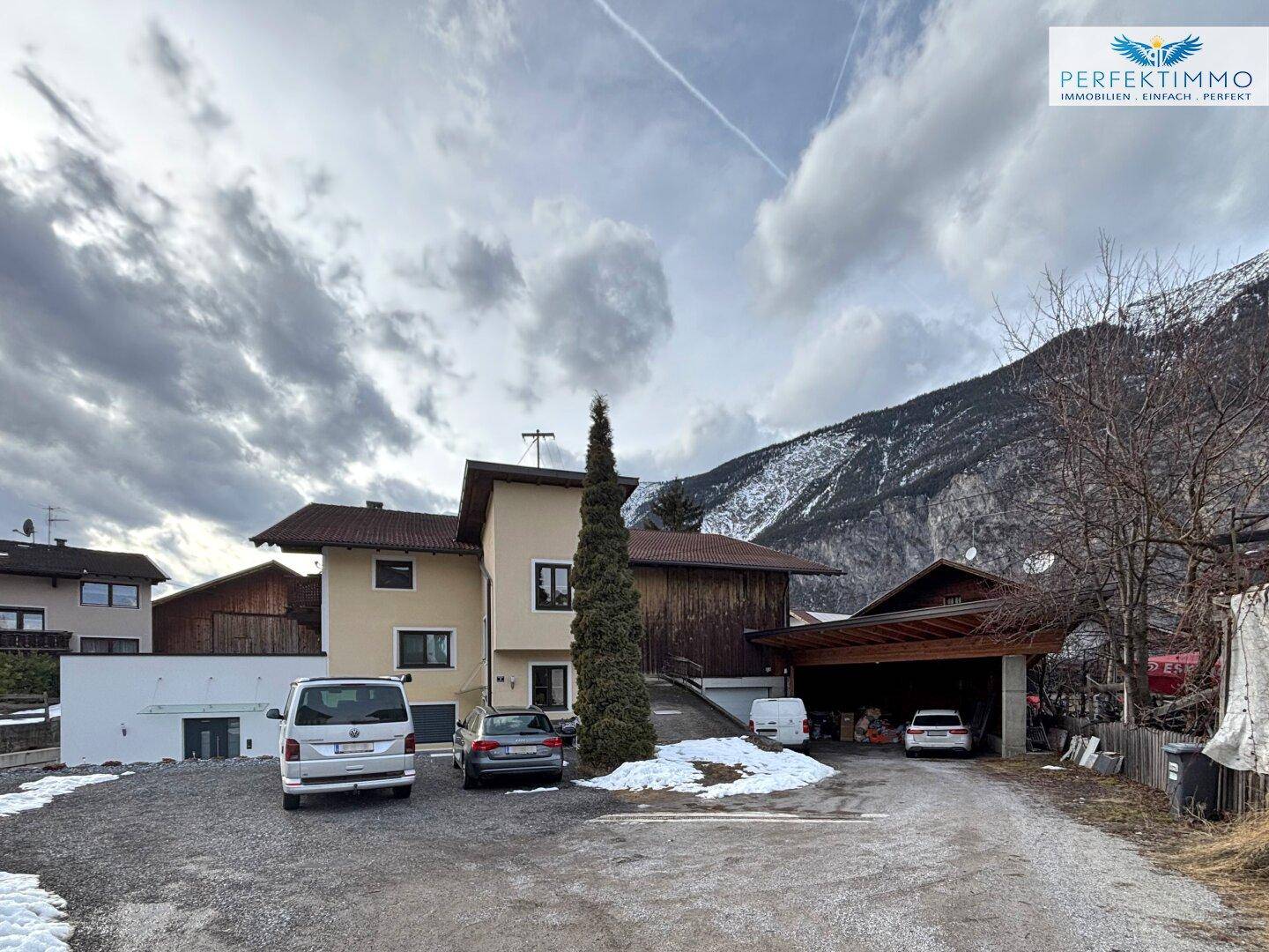 Ländliches Haus mit Bergblick und Parkplätzen im Vordergrund, umgeben von winterlicher Landschaft.