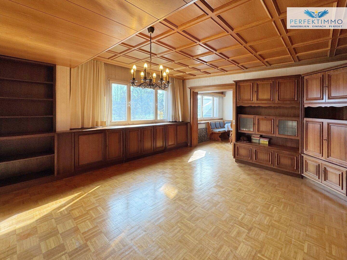 Geräumiges Wohnzimmer mit Holzkassettendecke, Parkettboden und großen Fenstern.