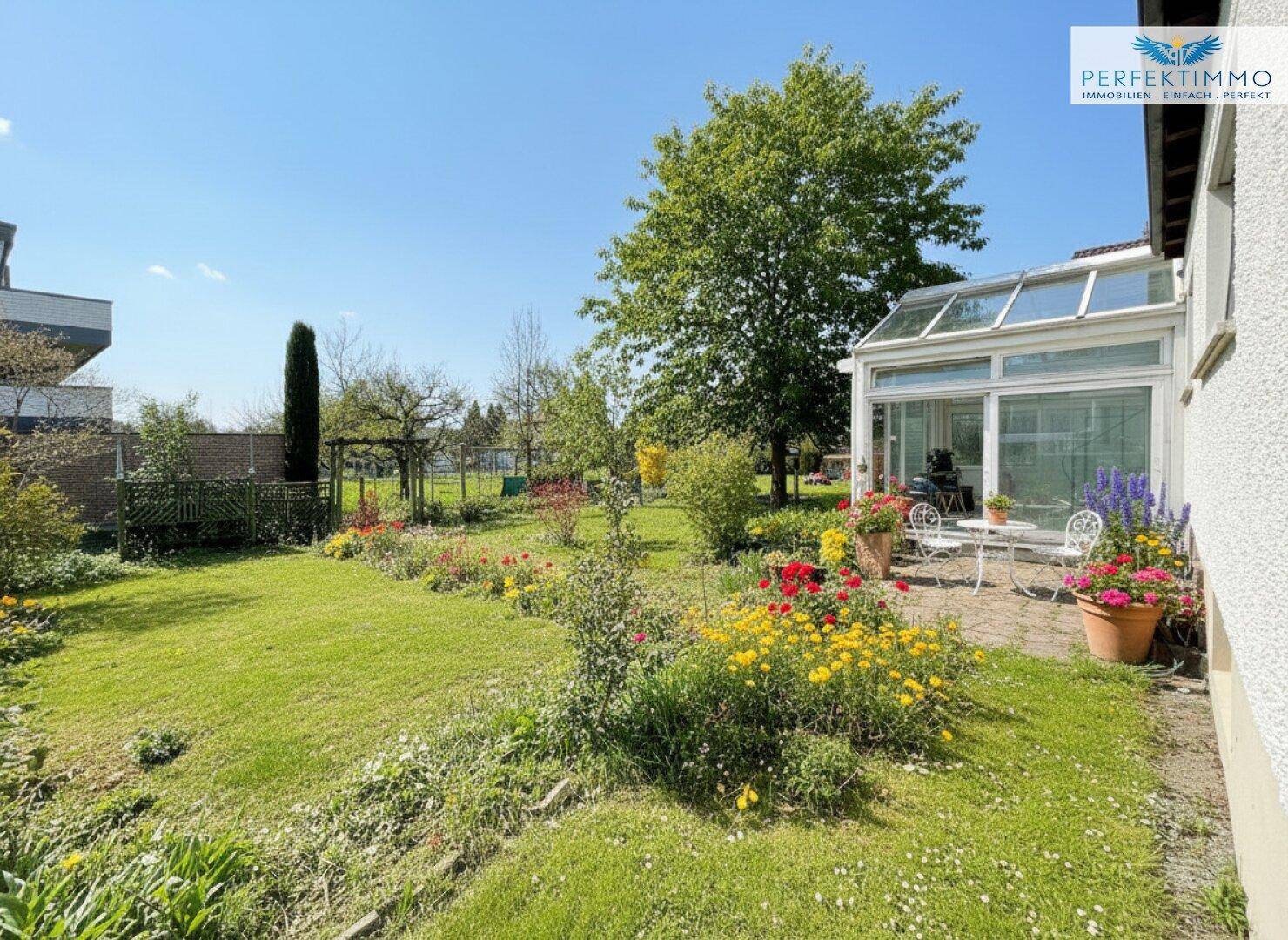 Großer, gepflegter Garten mit Rasenfläche, Blumenbeeten und einer Terrasse am Wintergarten.