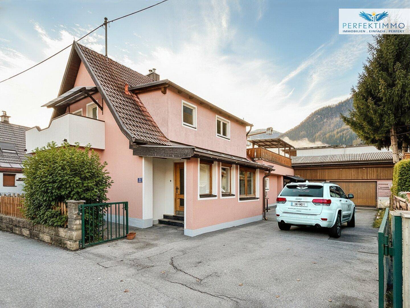 Rosa gestrichenes Einfamilienhaus mit Satteldach, Balkon und Stellplatz im Freien.