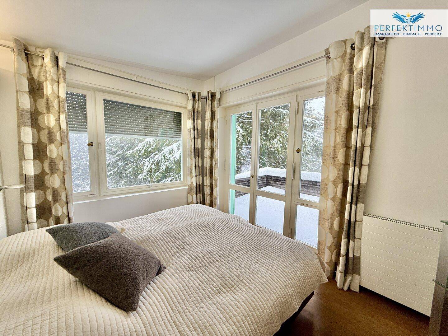 Schlafzimmer mit Doppelbett, Holzboden und Zugang zu einem Balkon mit Blick auf die verschneite Natur.