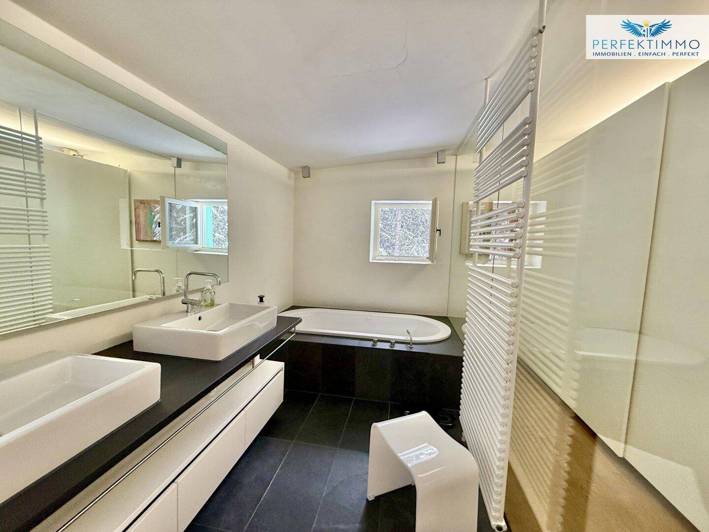 Helles Badezimmer mit Doppelwaschbecken, Badewanne und beheiztem Handtuchhalter, mit Fenster.