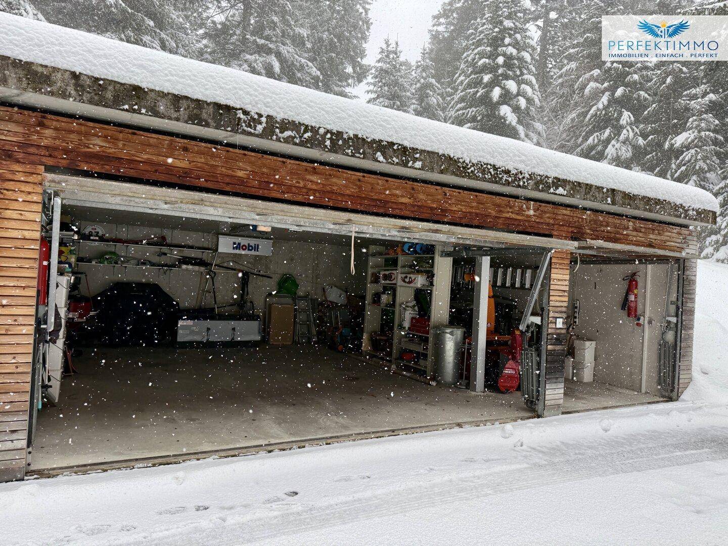 Offene Doppelgarage im Schnee, umgeben von verschneiten Tannenbäumen in winterlicher Landschaft.