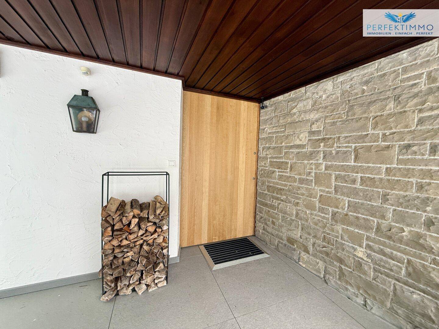 Überdachter Eingangsbereich mit Holztür, Natursteinwand und dekorativem Holzstapel für den Kamin.